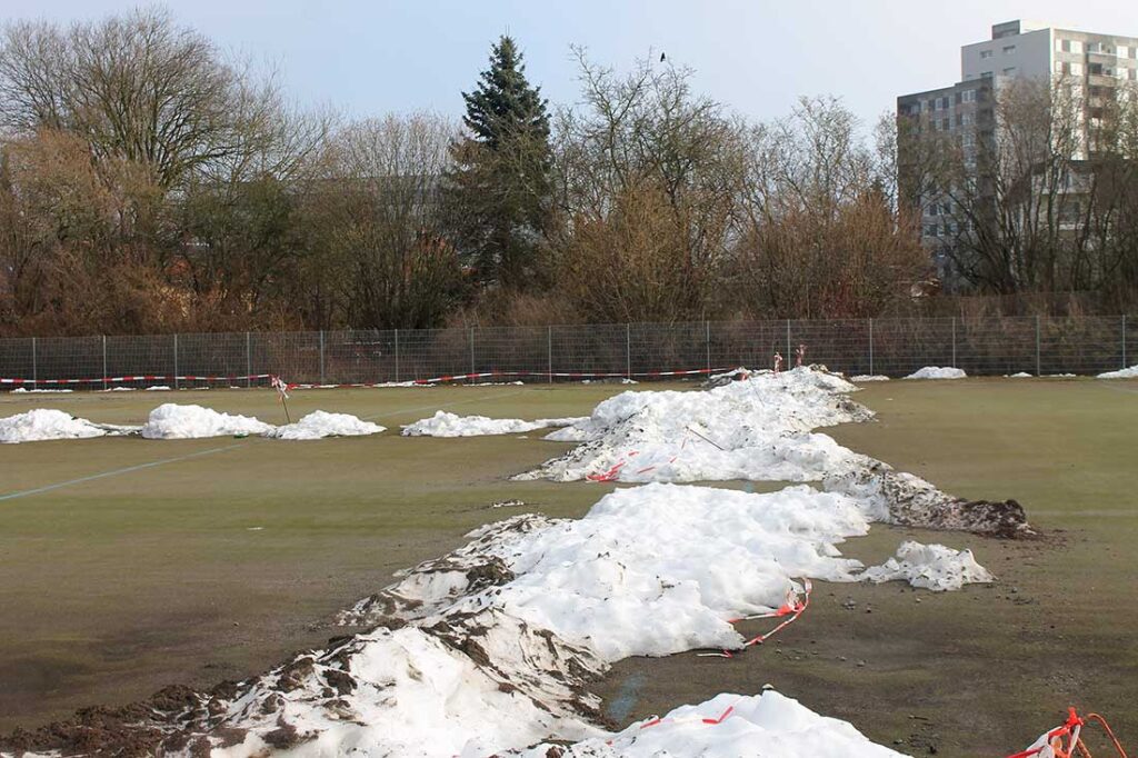 Unterschrift: Die traurigen Reste des "grünen" Fögeplatzes mit Schneeresten im Frühjahr 2026. Zuletzt diente er als Parkplatz. Blickrichtung und Standort ähnlich wie bei Bild 1.